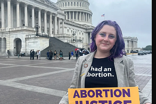A photo of Lizzie, a White woman with purple hair. She is wearing a T-shirt that reads “had an abortion,” and holding a sign that says “ABORTION JUSTICE NOW. The Capitol building is in the background.