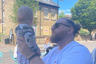 A man holding a baby. He has sunglasses on, and a white tshirt. The baby is wearing a camo long sleeved top and pink and white striped joggers.