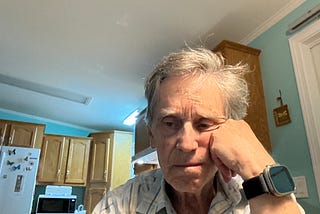 An older man with gray hair sits at a kitchen table, resting his face on his hand and appearing deep in thought or perhaps dejected. He is wearing a plaid shirt and a smartwatch. Behind him are wooden cabinets, a microwave, and a refrigerator with butterfly magnets. The lighting is bright, and the overall mood seems contemplative or sad.