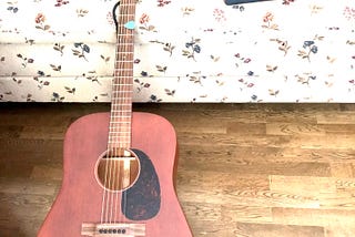 A picture of a mahogany wood acoustic Martin guitar, lying on a couch with a floral pattern. There’s a MacBook laptop opened up with the screen facing the other way.