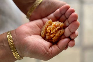 A brown colored sweet pudding is placed on the palm of cupped hands of a person who is also seen wearing golden bangles.