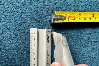 Blue carpet, ruler, tape measure, knife. The knife is being held as though the carpet is about to be cut.