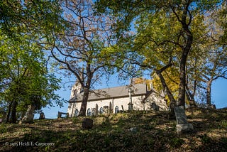 A view where one is looking up from the base of a hill at a classic church with steeple. Trees and graves line the hill.