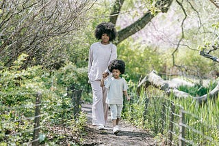 A Black woman, and her daughter, hold hands as they walk on a path at a park.