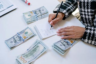 A person sitting with a notepad and pen and 4 wads of money writing accounts and money she spent and will save