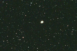 A bright oval shape in the middle of a dark star field. The oval is oriented with the long axis close to horizontal, slightly upward left to right. This is the Ring Nebula.