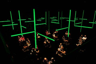 Visitors sit in the Donmar auditorium, 2 metres apart wearing headphones. A grid of green strip lights hangs above them.