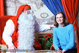 Santa with a long beard in red clothes holding his an arm on a shoulder of a brunette in a blue sweater. Both smiling at camera.