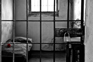 Black-and-white photo of a harsh prison cell on a remote island prison: thick metal bars in the foreground, a narrow bed with a single red rose on the mattress, and a small wooden table with a heavy padlock, illuminated by dim light through a high, barred window.