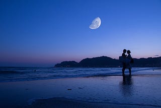color photograph of a couple standing on the beach at twilight with the moon aglow in the blue and fuschia sky