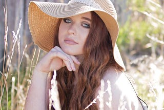 Young woman, brunette long hair, wears a floppy hat, surrounded by long grass, summer, countryside