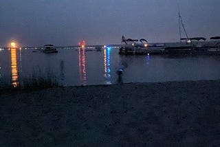 Distant fireworks shimmer over a large lake at twilight. In the foreground, a sandy beach with a patch of beach grass leads to shallow water with anchored and docked boats.