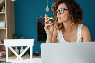 A woman with curly hair and glasses in front of her laptop. She’s keeping a pen and thinking.