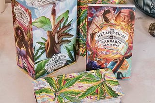 The Metaphysical Cannabis Oracle Deck and guidebook open on desk with cards placed in a stack in front of slip box cover. Photos of whimisically drawn cannabis leafs surround the cards.