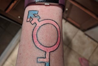 A picture of a tattoo, with a Transfeminine symbol combined with the Demigirl symbol in the colors of the Transfeminine Pride