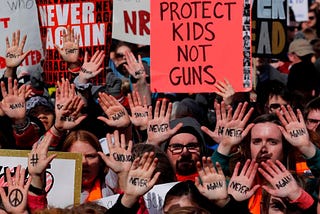 A crowd of people with their palms up that say “never again” and holding a sign that says protect kids not guns