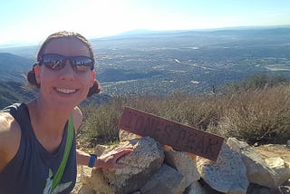 Smiling woman taking selfie with sign on pile of rocks that says, “Jones Peak” and view behind from top of mountain.