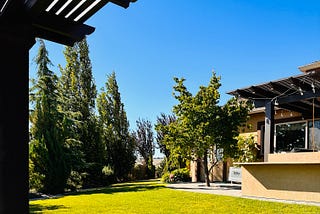 A backyard is shown with a lawn, pergola and tall trees, with blue sky in the background