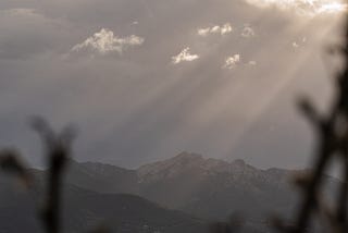 A dark mountain range with light breaking through dark clouds, in the foreground some blurry shapes of branches sticking out from the bottom