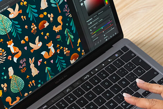 A close-up view of a graphic designer’s hands working on a laptop with design software open, displaying a colorful pattern featuring stylized forest animals and plants on a dark background. The screen shows a vibrant, detailed illustration with various tools and color palettes, indicating the digital creation process of children’s pattern design.