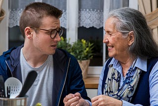 A young man with glasses sits at a kitchen table with a smiling older woman in blue with gray hair