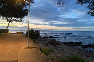 A coastal promenade in france, lit by lampposts in the early morning. The sky is just getting lighter with some clouds scudding the sky. The sea and some rocks and a corner of beach can be seen, with gentle waves coming in and trees lining some of the path