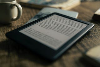 A black Kindle is set on a tabletop next to a white coffee mug, and resting on top of another book of some sort.