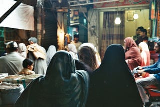 A market in Lahore, Pakistan. Women and men dressed in traditional garb walk through a darkly lit market.