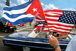 A man in a dark blue car is holding an American flag while the person closer to the camera is waving a Cuban flag