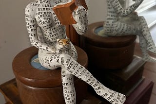 The photo shows a paper-mache figurine. It is a seated woman holding an opened book in her left hand and a small cat perched on her lap. Her right leg is crossed over her left one.