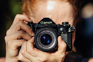 Photo of a woman holding a 1970s-style film camera to her face to take a photo.