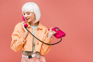 young woman in white-blonde wig wearing an orange jacket talking on a hot pink corded phone on dusty rose background