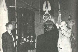 A man gives a Roman Salute to a flag with an eagle perched on a wreath. In the wreath are three tools.