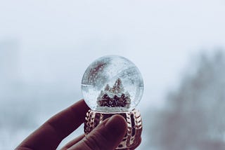 A hand holding a glass snow globe with a house inside.