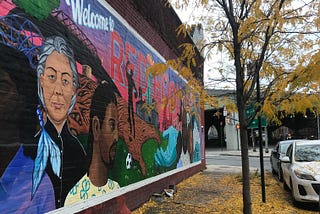 Colorful wall art depicting diverse people lives on a Red Hook building next to black tree with falling yellow leaves