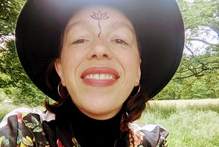 Gillian Torres sits in Sefton Park smiling, in a floral jacket and black hat, with a lotus flower drawn on her third eye.
