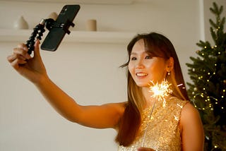 An Asian woman with long dark hair and bangs holds a sparkler in one hand and her black cell phone on a selfie stick in the other. She is wearing a gold dress and smiling as she stands in front of a twinkling Christmas tree with white lights.