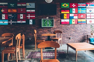 Mesas em um restaurante, com uma parede cheia de relógios ao fundo, cada relógio tendo a bandeira de um país.