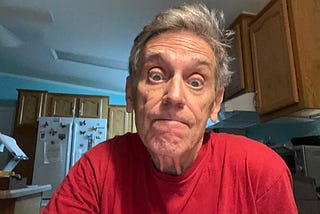 A man with gray hair wearing a red shirt looks directly at the camera shrugging with a raised-eyebrow expression.