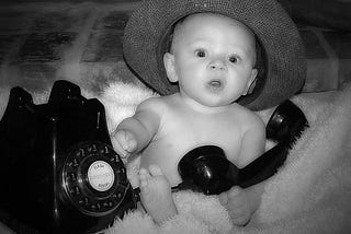 infant in a sunhat holding  an old-school rotary telephone