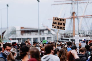 A crowd protesting with a sign held up saying “Racism will not be tolerated”.