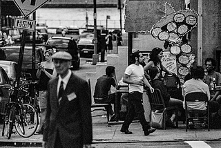 Black and white photo of a street corner in DUMBO, Brooklyn. An older man in a cap is crossing the street, while a woman behind him stands looking at her mobile device. People sit outside a Mexican restaurant as another pedestrian (or perhaps restaurant server) passes by.