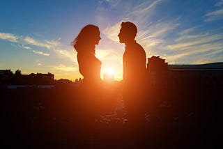 A couple stands silhouetted against a setting sun.