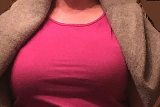 closeup of a very large chest straining the material of a pink shirt in multiple lines across the front and up and down in the shoulders to cover the collarbone; a large grey sweater drapes over the pale white shoulders.