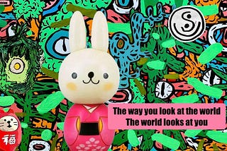 Colorful illustration by artist Francois Top featuring a white rabbit doll dressed in a pink kimono standing in front of a vibrant mural filled with abstract faces, eyes, plants, and colorful shapes. A small red Japanese-style figurine appears in the lower left corner, and a circular symbol with the letter “S” appears in the upper right. A pink text box reads: “The way you look at the world. The world looks at you.”