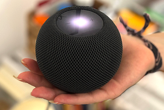 Na imagem, uma mão segurando um Home Pod Mini da Apple, um dispositivo redondo de cor escura com uma luz brilhante no meio. Uma interface conversacional que permite com que as pessoas possam interagir com o ambiente e utilizar inteligências computacionais a partir de comandos de voz.
