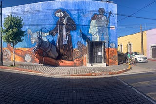 Detail of a mural in Cholula, Puebla, Mexico, artist unkown. Photo by Jim Latham. A priest palms a gem while a coquistador stabs an Indigenous man. Another Spaniard with a crossbow menaces an Indigenous woman, possibly La Malinche.