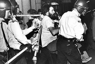 During a riot, a resisting man with a beard is being carried by four police officers.