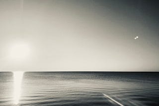 A symbolic monochromatic rendering of a photograph, with an almost invisible and forgotten sun, setting over the horizon of seemingly calm and undistrubed waters.