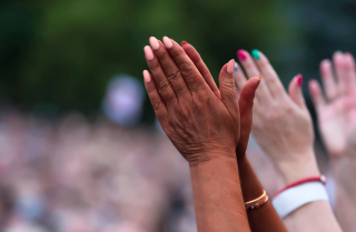 Image is of Black and white people’s hands, clapping in a crowd at an outdoor rally.